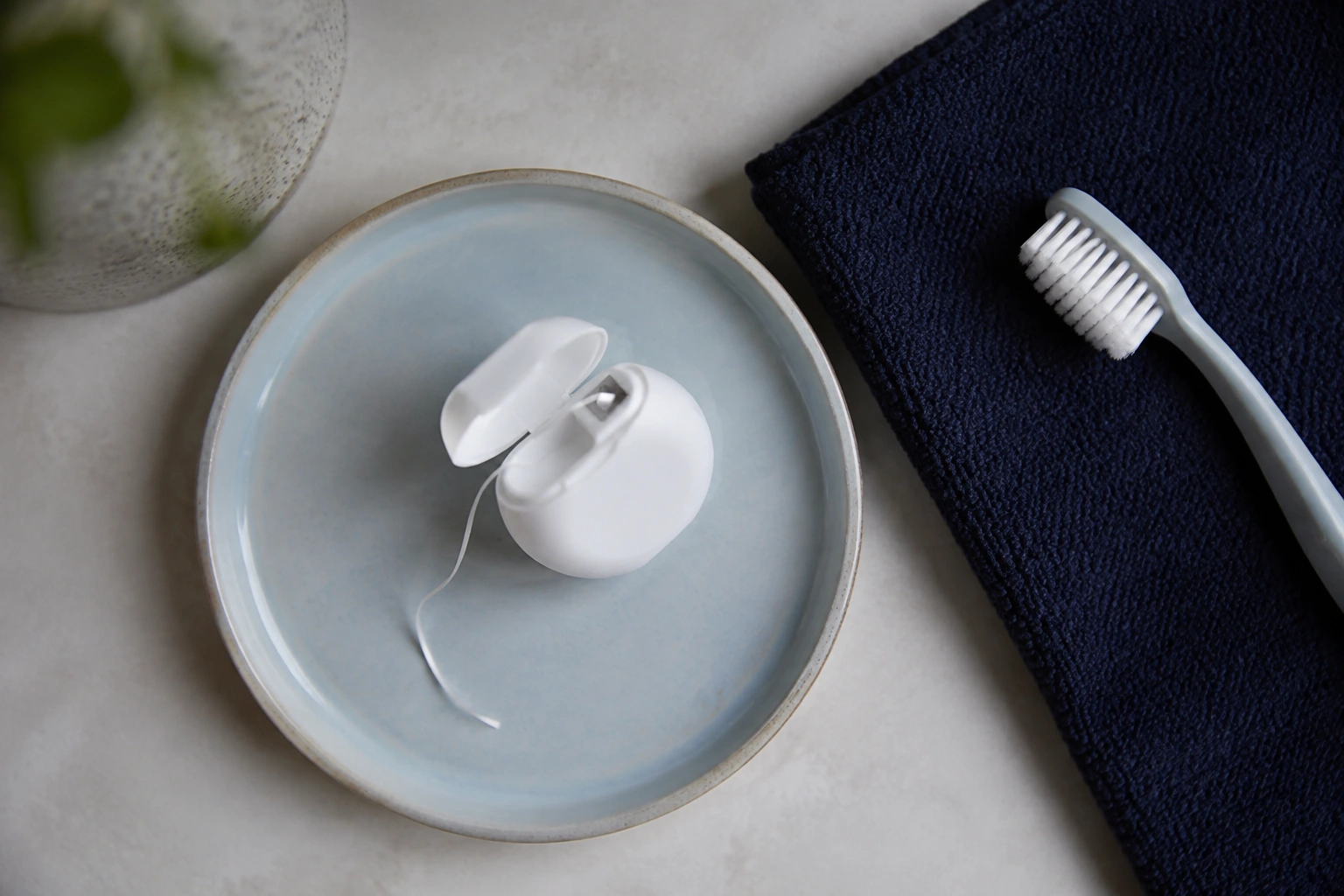 An open container of floss with a short single strand on a pale blue ceramic dish beside a soft toothbrush and folded navy washcloth.