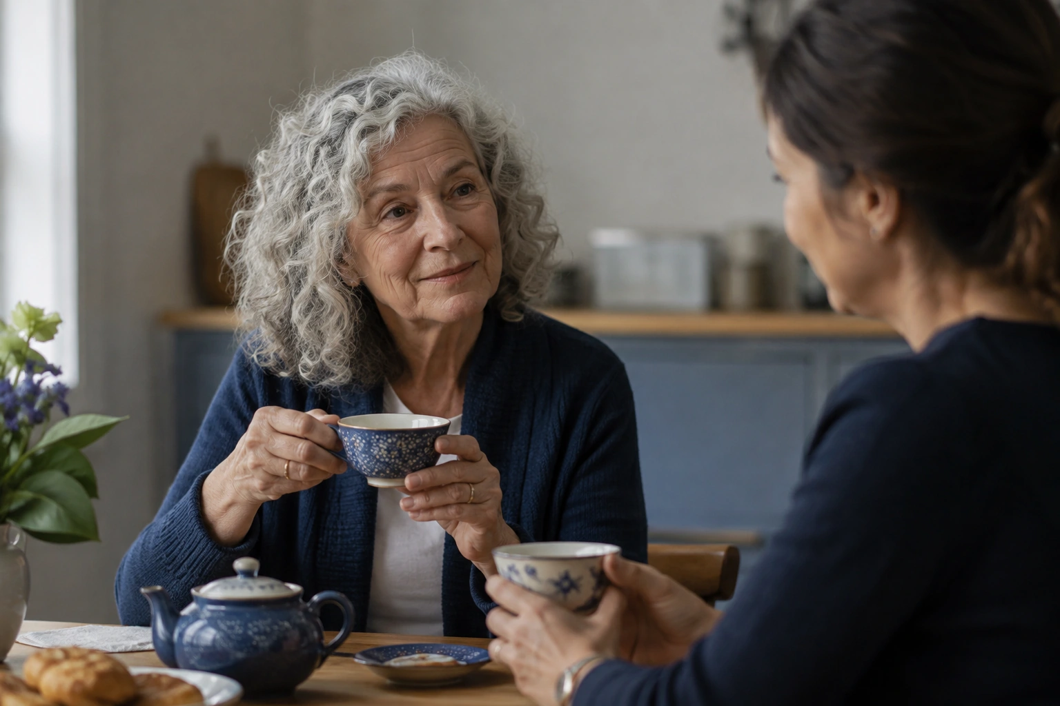 An older woman with silver curls sharing tea at a kitchen table.