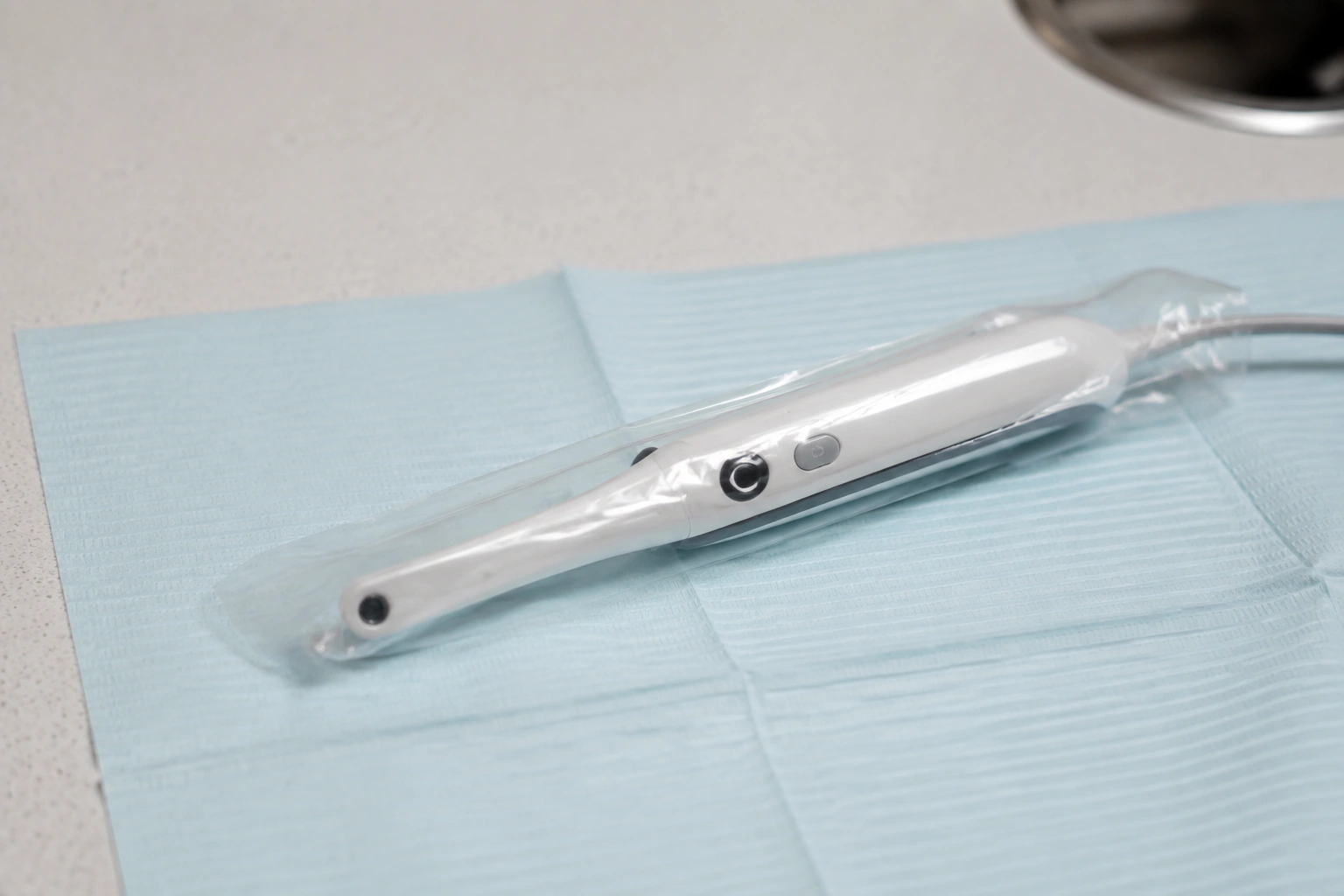 Macro view of a slim intraoral camera handpiece in a transparent protective sleeve lying on folded pale-blue dental paper.