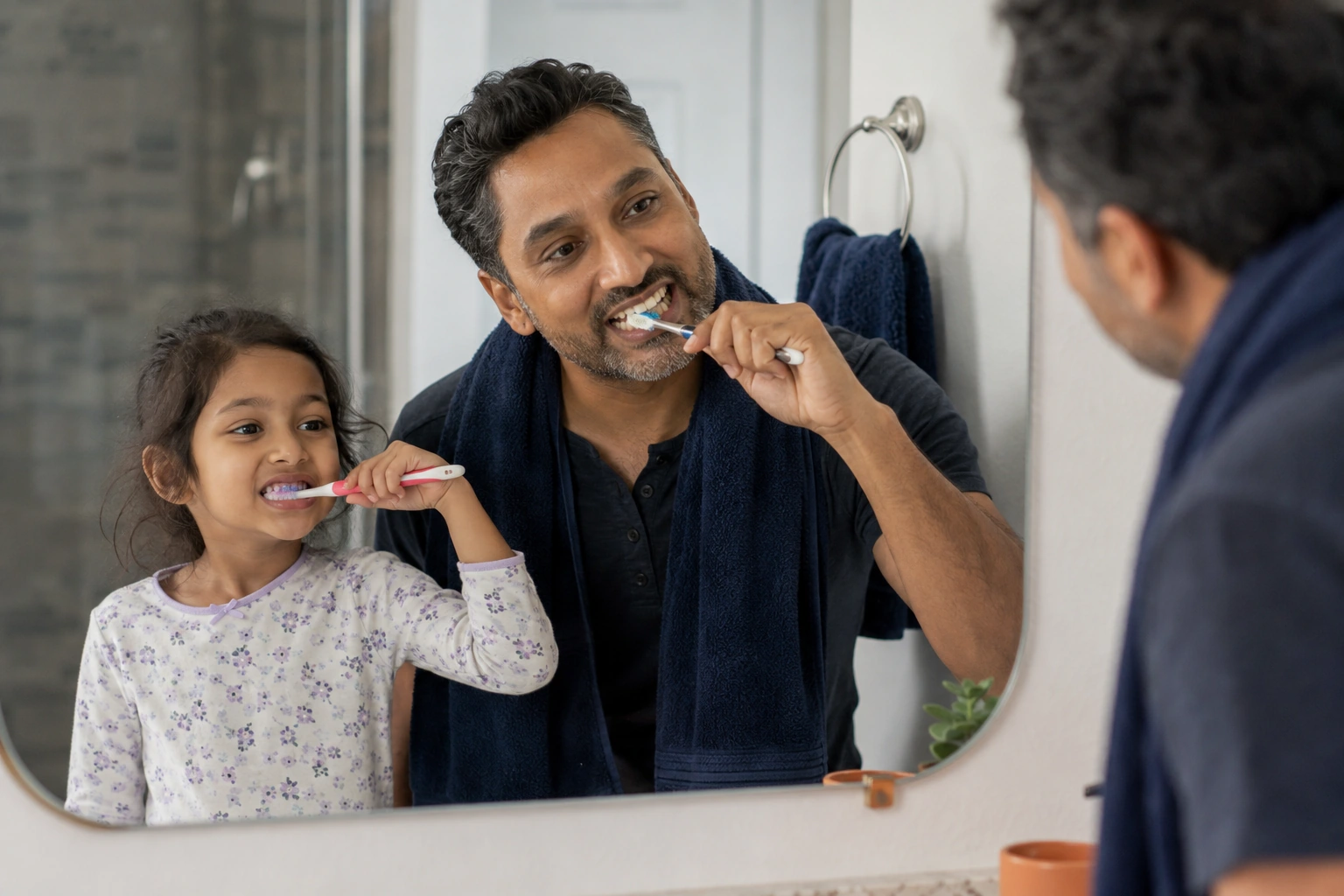 A South Asian father and his seven-year-old daughter brushing their teeth side by side at home.