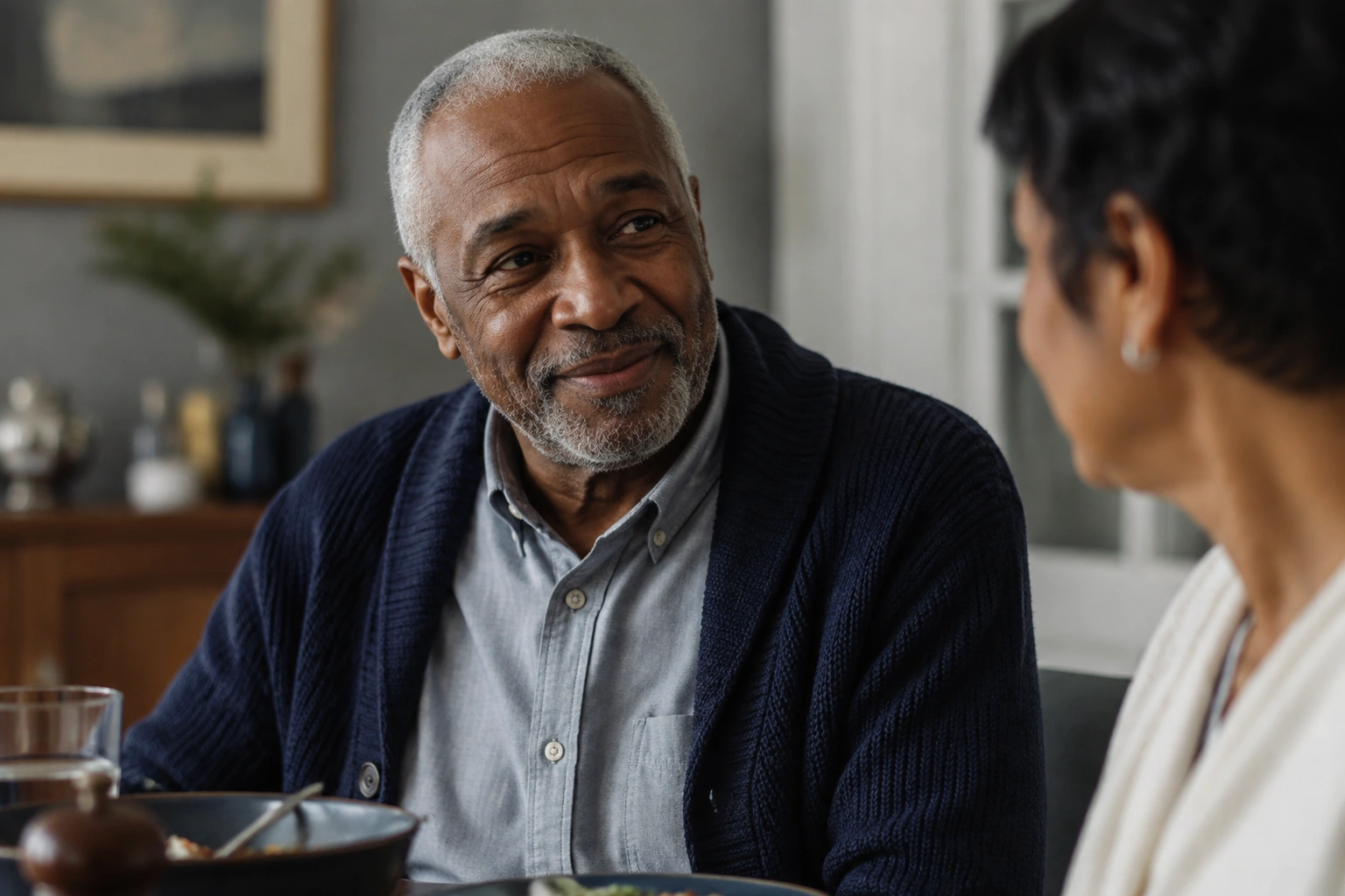An older Black man smiling naturally over a home lunch conversation.