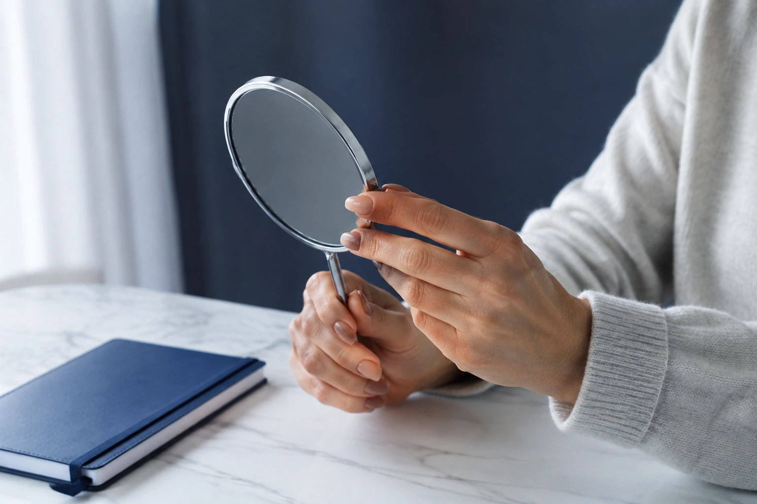 A woman’s hands holding a small hand mirror at a consultation table beside a closed notebook.