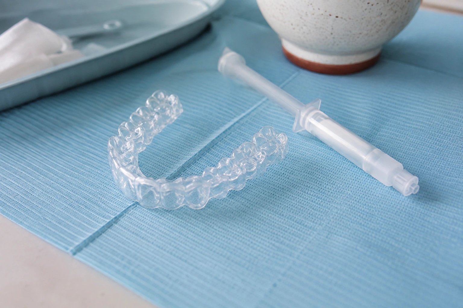 A clear custom whitening tray resting on blue dental paper beside an unlabeled capped gel syringe.